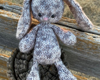 May include: A knitted gray and white bunny rabbit with pink nose and black eyes sitting on a wooden fence post.