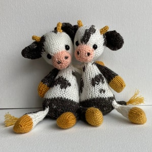 May include: Two knitted plush cow toys with black and white spots and yellow accents. The cows are facing each other and appear to be hugging.