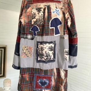 May include: A long-sleeved button-down shirt featuring a patchwork design of plaid, striped, and floral fabrics. The garment includes bleached sections and various fabric appliqu&eacute;s, including two dark blue mushroom shapes and a central heart patch.
