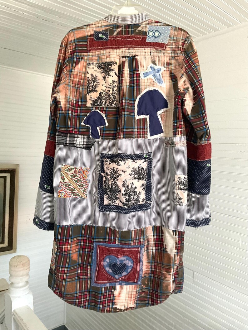 May include: A long-sleeved button-down shirt featuring a patchwork design of plaid, striped, and floral fabrics. The garment includes bleached sections and various fabric appliqu&eacute;s, including two dark blue mushroom shapes and a central heart patch.