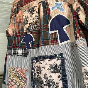 May include: A long-sleeve button-down shirt featuring a patchwork design of plaid, striped, and toile fabric. The garment includes appliqued dark blue mushroom shapes, a light blue bird patch, and various rectangular fabric squares stitched on.