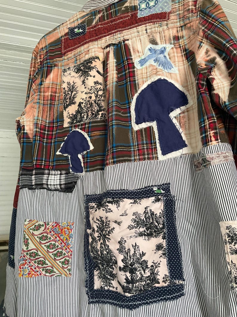May include: A long-sleeve button-down shirt featuring a patchwork design of plaid, striped, and toile fabric. The garment includes appliqued dark blue mushroom shapes, a light blue bird patch, and various rectangular fabric squares stitched on.