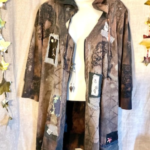 May include: A long-sleeve hooded zip-up jacket in shades of brown and gray with a mottled, distressed finish. The garment features multiple rectangular fabric patches with various patterns, including stripes and gear motifs, and a dangling metal pendant.