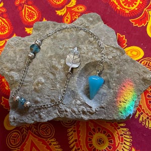 May include: A silver chain pendulum with a blue crystal point and a silver leaf charm. The chain has blue beads and rests on a gray stone. The background is a red and yellow patterned fabric.