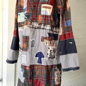 May include: A long-sleeved shirt featuring a patchwork design of red, blue, and green plaid fabric combined with black and white pinstriped panels. The garment includes various fabric appliqu&eacute;s, including a blue mushroom shape and a toile print.