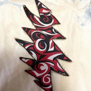 May include: A cream-colored fabric with a large, stylized lightning bolt applique. The bolt features a red, black, and white swirling pattern, outlined with black stitching. The fabric has a light blue tie-dye effect.