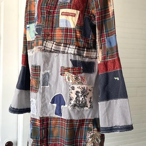 May include: A long-sleeved shirt featuring a patchwork design of red, green, and blue plaid fabric combined with sections of black and white pinstriped fabric. The garment includes various fabric patches in floral, polka dot, and toile patterns.