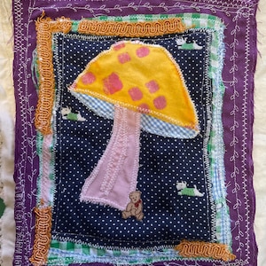 May include: A rectangular fabric wall hanging featuring a central dark blue polka dot panel with a large yellow mushroom with pink spots, a small brown teddy bear, and three small white dogs. The piece is layered with purple, green gingham, and orange trim.