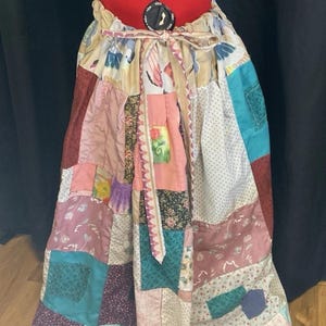 May include: A patchwork skirt with a variety of fabric patterns and colors, including floral, polka dot, and solid patches. The skirt is tied at the waist with a matching fabric tie. The skirt is displayed on a red dress form.