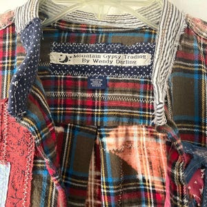 May include: A patchwork shirt featuring various plaid patterns in red, blue, and brown. The collar has a striped fabric trim. A label reads Mountain Gypsy Trading By Wendy Darling, with a size M tag indicating 100% cotton, made in Nicaragua.