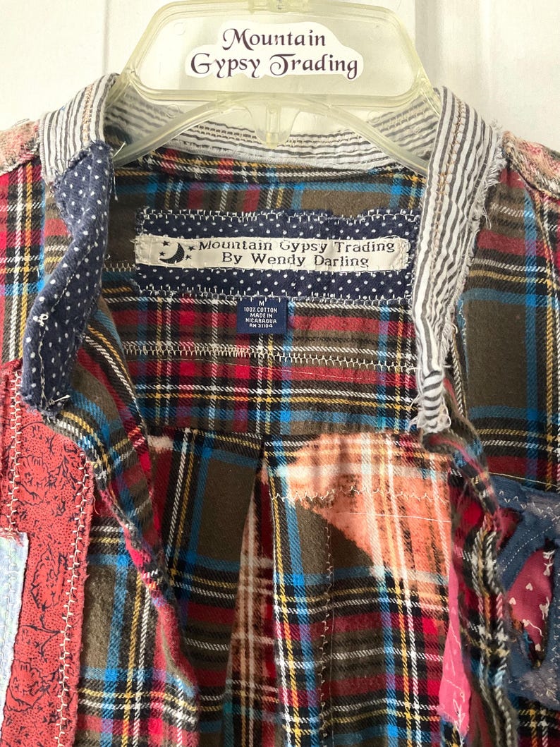 May include: A patchwork shirt featuring various plaid patterns in red, blue, and brown. The collar has a striped fabric trim. A label reads Mountain Gypsy Trading By Wendy Darling, with a size M tag indicating 100% cotton, made in Nicaragua.