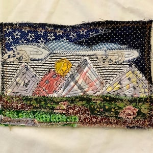 May include: A rectangular fabric collage featuring a night sky with white stars on a dark blue background, stylized clouds, a yellow sun, and four triangular mountain peaks made of various patterned fabrics above a green landscape.