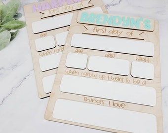 May include: Two wooden signs with blank spaces for writing. The top sign says "HARPER'S last day of" in pink and white letters. The bottom sign says "BRENDYN'S first day of" in blue and white letters.