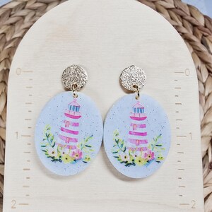 May include: A pair of oval-shaped earrings featuring a lighthouse design. The earrings have a light blue background with a glitter effect. The lighthouse is pink and white, with a gold top. Floral accents are at the base, and the earring posts are gold.