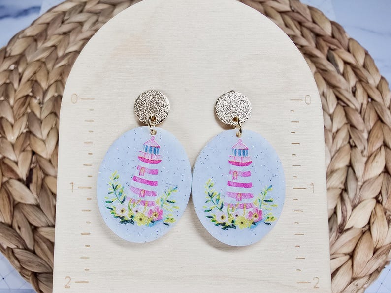 May include: A pair of oval-shaped earrings featuring a lighthouse design. The earrings have a light blue background with a glitter effect. The lighthouse is pink and white, with a gold top. Floral accents are at the base, and the earring posts are gold.