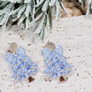 May include: A pair of Christmas tree earrings. Each earring features a blue and white floral design with a bow at the top and a brown trunk. The earrings have a sparkly, silver-colored stud.