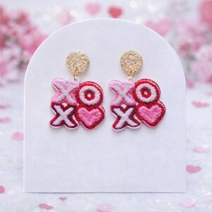 May include: A pair of Valentine's Day earrings. Each earring features the letters "XO" in a pink and red embroidered design, with a heart shape. The earrings have gold-colored stud posts and are displayed on a white stand.