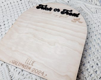 May include: A light wood sign with a rounded top and the text "Trick or Treat! Smell my feet!" in black lettering. The sign also has the text "Ellie Halloween 2024" in brown lettering.