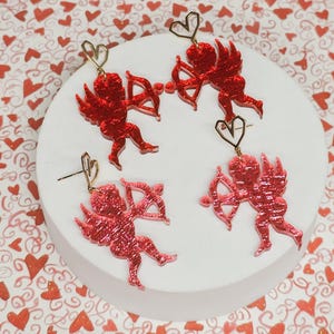 Puede incluir: Cuatro pares de pendientes de cupido de purpurina roja con detalles dorados. Cada pendiente presenta un cupido con alas, sosteniendo una flecha y un corazón.