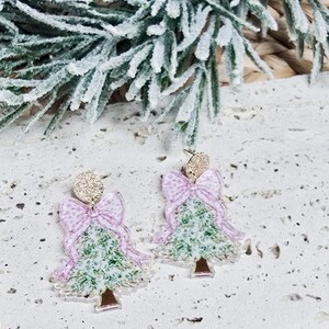 May include: A pair of Christmas tree earrings. Each earring features a green tree design with a pink bow and a gold glitter top. The earrings are set against a textured white surface with frosted greenery.