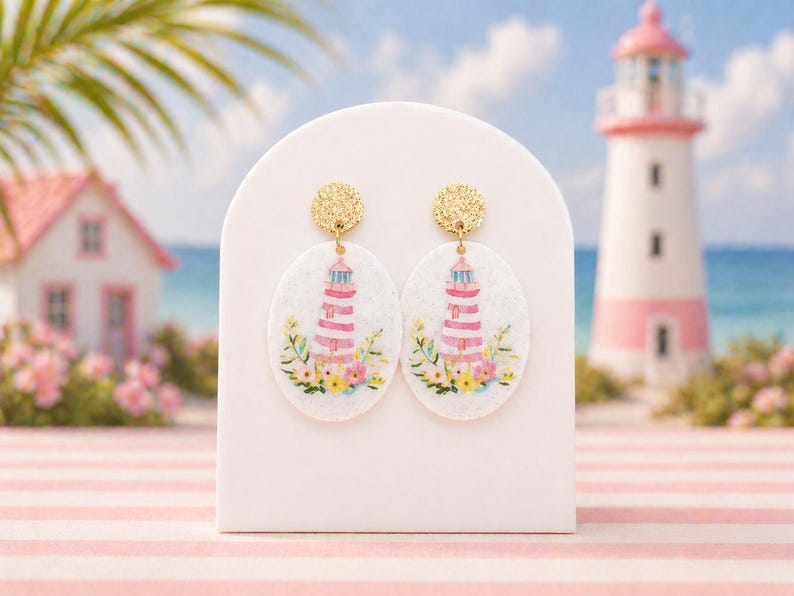 May include: A pair of gold-toned, textured stud earrings with oval pendants. Each pendant features a watercolor illustration of a lighthouse with pink and white stripes, surrounded by flowers. The earrings are displayed on a white stand, with a lighthouse and ocean in the background.