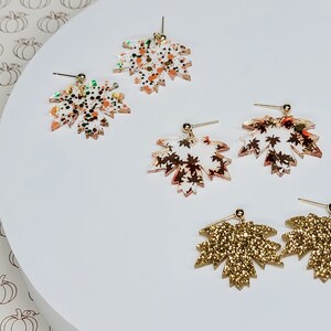May include: A set of six gold and white leaf-shaped earrings. The earrings are made of acrylic and have a glitter finish. The leaves are different sizes and shapes, and some have a speckled pattern.