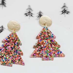May include: A pair of festive Christmas tree earrings. Each earring features a gold-colored stud and a tree-shaped pendant filled with colorful glitter. The earrings are set against a white background with a subtle tree pattern.