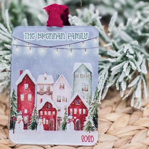 Puede incluir: Adorno navideño rectangular con una escena de pueblo invernal, con casas cubiertas de nieve en rojo, blanco y verde. El texto "THE BRENNAN FAMILY" está en la parte superior, con "2025" en la inferior. Una cinta roja está adjunta para colgar.