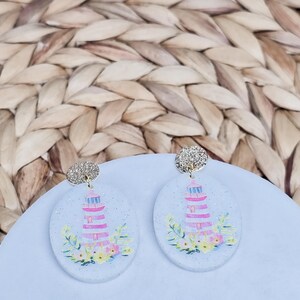 May include: A pair of oval-shaped earrings featuring a lighthouse design. The earrings have a glittery silver top and a light blue base with a pink and white lighthouse illustration, surrounded by colorful flowers and greenery.