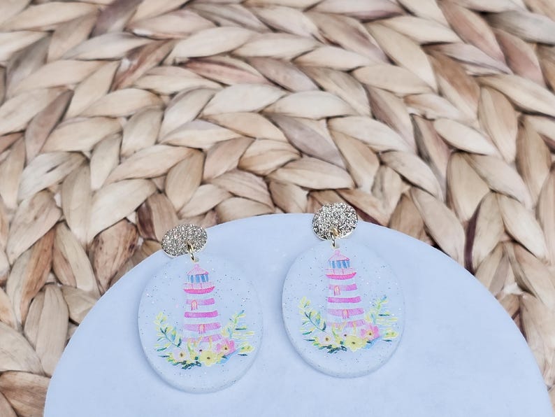May include: A pair of oval-shaped earrings featuring a lighthouse design. The earrings have a glittery silver top and a light blue base with a pink and white lighthouse illustration, surrounded by colorful flowers and greenery.
