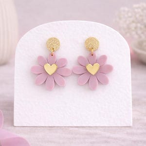 Puede incluir: Un par de pendientes de margarita lavanda con centros de corazón dorados y detalles superiores de purpurina dorada. Los pendientes se exhiben sobre una superficie blanca, mostrando un diseño de joyería floral.