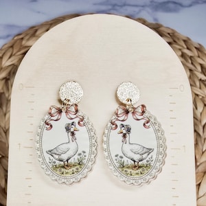 Puede incluir: Un par de pendientes colgantes ovalados con una oca blanca con capota, enmarcados por un borde festoneado y una cinta rosa. Los pendientes est&aacute;n unidos a tachuelas circulares doradas con textura y miden 3,8 cm de altura.