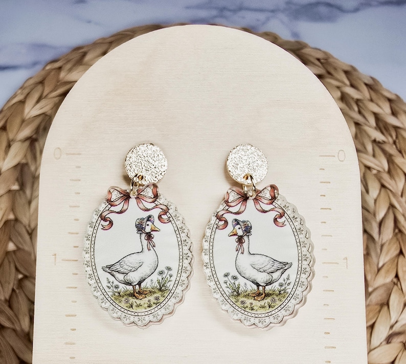 Puede incluir: Un par de pendientes colgantes ovalados con una oca blanca con capota, enmarcados por un borde festoneado y una cinta rosa. Los pendientes est&aacute;n unidos a tachuelas circulares doradas con textura y miden 3,8 cm de altura.