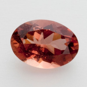 May include: An oval-shaped gemstone with a faceted surface. The gem has a warm, reddish-orange color with internal patterns and inclusions. The gemstone is set against a plain white background, highlighting its color and cut.