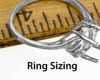 Puede incluir: Primer plano de un engaste de anillo plateado con un diseño de garras, listo para una piedra preciosa. El anillo está cerca de una regla de madera, con el texto "Ring Sizing" en negro.