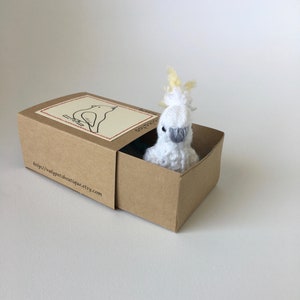 K&ouml;nnte beinhalten: Ein handgefertigtes, wei&szlig;-graues geh&auml;keltes Kakadu-Vogelspielzeug schaut aus einer kleinen, offenen braunen Pappschachtel. Die Schachtel hat ein Etikett mit einer Vogelillustration und dem Text "valypetsboutique.etsy.com". Der Vogel hat einen gelben Schopf.