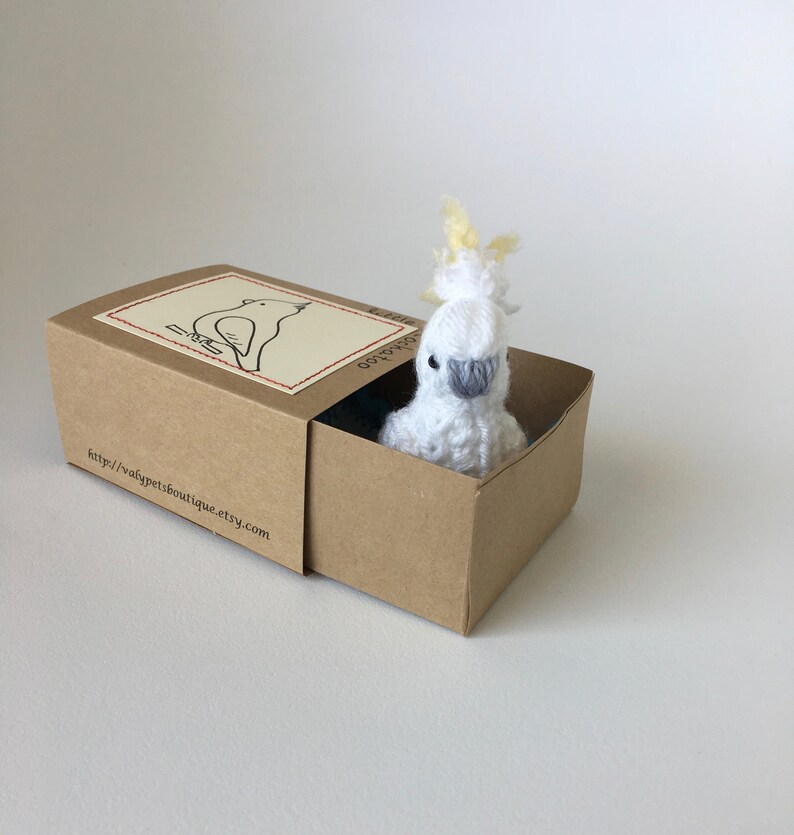 K&ouml;nnte beinhalten: Ein handgefertigtes, wei&szlig;-graues geh&auml;keltes Kakadu-Vogelspielzeug schaut aus einer kleinen, offenen braunen Pappschachtel. Die Schachtel hat ein Etikett mit einer Vogelillustration und dem Text "valypetsboutique.etsy.com". Der Vogel hat einen gelben Schopf.