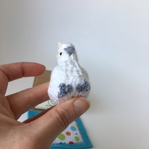 K&ouml;nnte beinhalten: Ein kleiner, wei&szlig; geh&auml;kelter Vogel mit grauen Akzenten auf Fl&uuml;geln und F&uuml;&szlig;en. Der Vogel hat einen gelben Schnabel und wird in einer Hand gehalten. Eine kleine blaue Filzmatte mit einem bunten Tupfenmuster befindet sich im Hintergrund.