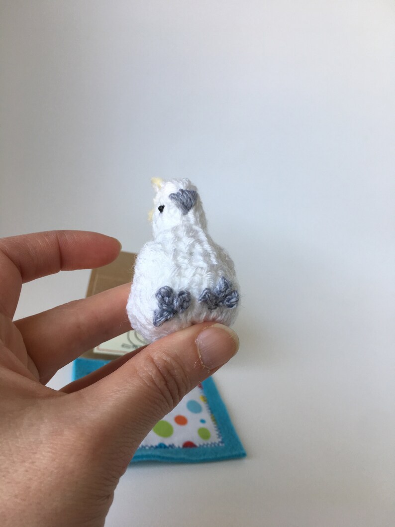 K&ouml;nnte beinhalten: Ein kleiner, wei&szlig; geh&auml;kelter Vogel mit grauen Akzenten auf Fl&uuml;geln und F&uuml;&szlig;en. Der Vogel hat einen gelben Schnabel und wird in einer Hand gehalten. Eine kleine blaue Filzmatte mit einem bunten Tupfenmuster befindet sich im Hintergrund.