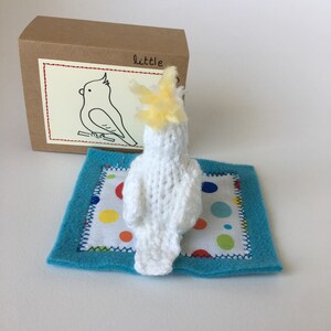 K&ouml;nnte beinhalten: Ein handgefertigter, wei&szlig;er, gestrickter Kakadu mit gelbem Kamm, der auf einer blauen Filzmatte mit einem Tupfenmuster ruht. Eine braune Pappschachtel mit einer Kakadu-Zeichnung und dem Wort "little" im Hintergrund.