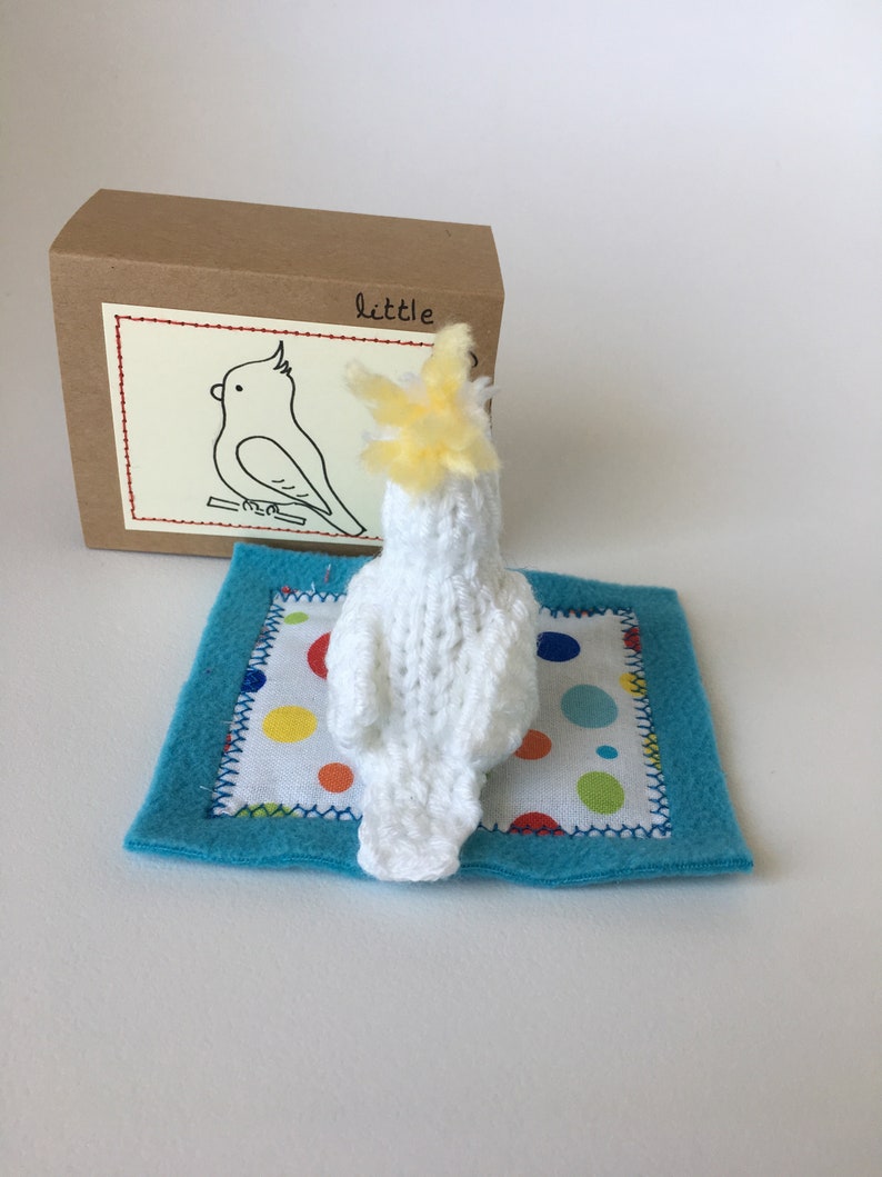 K&ouml;nnte beinhalten: Ein handgefertigter, wei&szlig;er, gestrickter Kakadu mit gelbem Kamm, der auf einer blauen Filzmatte mit einem Tupfenmuster ruht. Eine braune Pappschachtel mit einer Kakadu-Zeichnung und dem Wort "little" im Hintergrund.