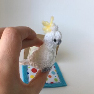 K&ouml;nnte beinhalten: Handgefertigte Figur eines wei&szlig;en und gelben Kakadus. Der Vogel hat eine gelbe Federhaube, graue F&uuml;&szlig;e und ein kleines schwarzes Auge. Er sitzt auf einer kleinen, bunten, quadratischen Matte mit blauem Rand. Der Vogel ist aus Garn.