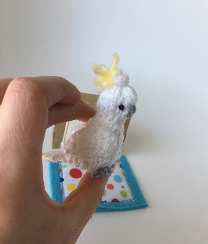 K&ouml;nnte beinhalten: Handgefertigte Figur eines wei&szlig;en und gelben Kakadus. Der Vogel hat eine gelbe Federhaube, graue F&uuml;&szlig;e und ein kleines schwarzes Auge. Er sitzt auf einer kleinen, bunten, quadratischen Matte mit blauem Rand. Der Vogel ist aus Garn.