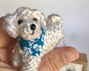 Könnte beinhalten: Handgefertigte weiße Häkelhund-Figur mit blauem Bandana mit Blumenmuster. Der kleine Hund hat schwarze Augen und eine schwarze Nase. Die Figur wird in einer Hand gehalten, mit einer kleinen Illustration und gemustertem Stoff im Hintergrund.