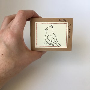 K&ouml;nnte beinhalten: Eine kleine, quadratische, braune Karte mit einer handgezeichneten Illustration eines Kakadus. Der Vogel ist mit schwarzer Tinte auf einem Ast dargestellt, mit den Worten "little Cockatoo". Ein roter, gen&auml;hter Rand umrahmt das Bild.