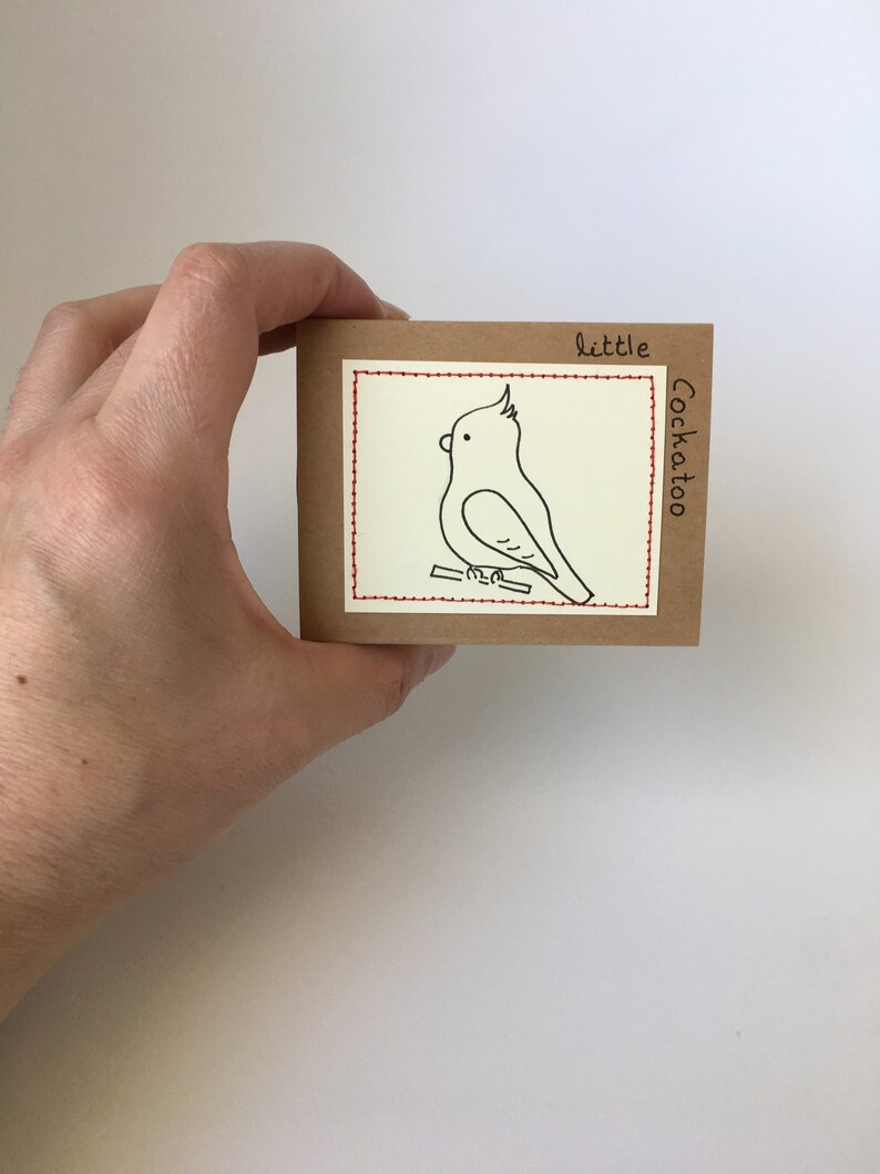 K&ouml;nnte beinhalten: Eine kleine, quadratische, braune Karte mit einer handgezeichneten Illustration eines Kakadus. Der Vogel ist mit schwarzer Tinte auf einem Ast dargestellt, mit den Worten "little Cockatoo". Ein roter, gen&auml;hter Rand umrahmt das Bild.