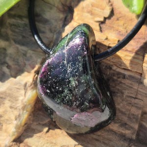Può includere: Un ciondolo in pietra preziosa lucidata con un cordino in pelle nera. La pietra presenta tonalità verde scuro, nere e rosa, con una finitura lucida. Il ciondolo è esposto su una superficie di legno.