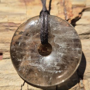 Könnte beinhalten: Ein klarer, donut-förmiger Anhänger mit einem Loch in der Mitte, der an einer dunkelgrauen Kordel hängt. Der Anhänger hat ein rauchiges, durchscheinendes Aussehen mit inneren Mustern. Der Hintergrund ist eine hellbraune, strukturierte Oberfläche.