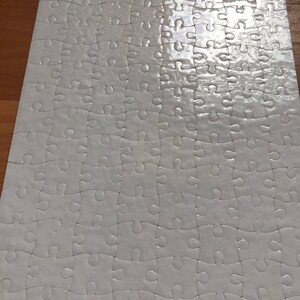 May include: A completed white jigsaw puzzle with interlocking pieces. The puzzle is rectangular and appears to be on a wooden surface. The pieces have a slight sheen, reflecting light.