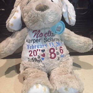 May include: A beige plush dog with white ears and paws. The dog is wearing a white shirt with the text "Noelle Harper Sciarre February 13, 2017 20" 8lb 6oz 7:59am" embroidered in pink, blue, and red.